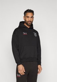 Black hoodie with a front pocket, ribbed cuffs and hem, featuring white and red embroidered logos on the chest, made of soft fabric.