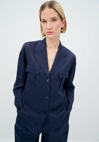 Blazer in lino blu navy con colletto a scollo, due tasche sul petto e tre bottoni frontali; texture liscia con vestibilità comoda.