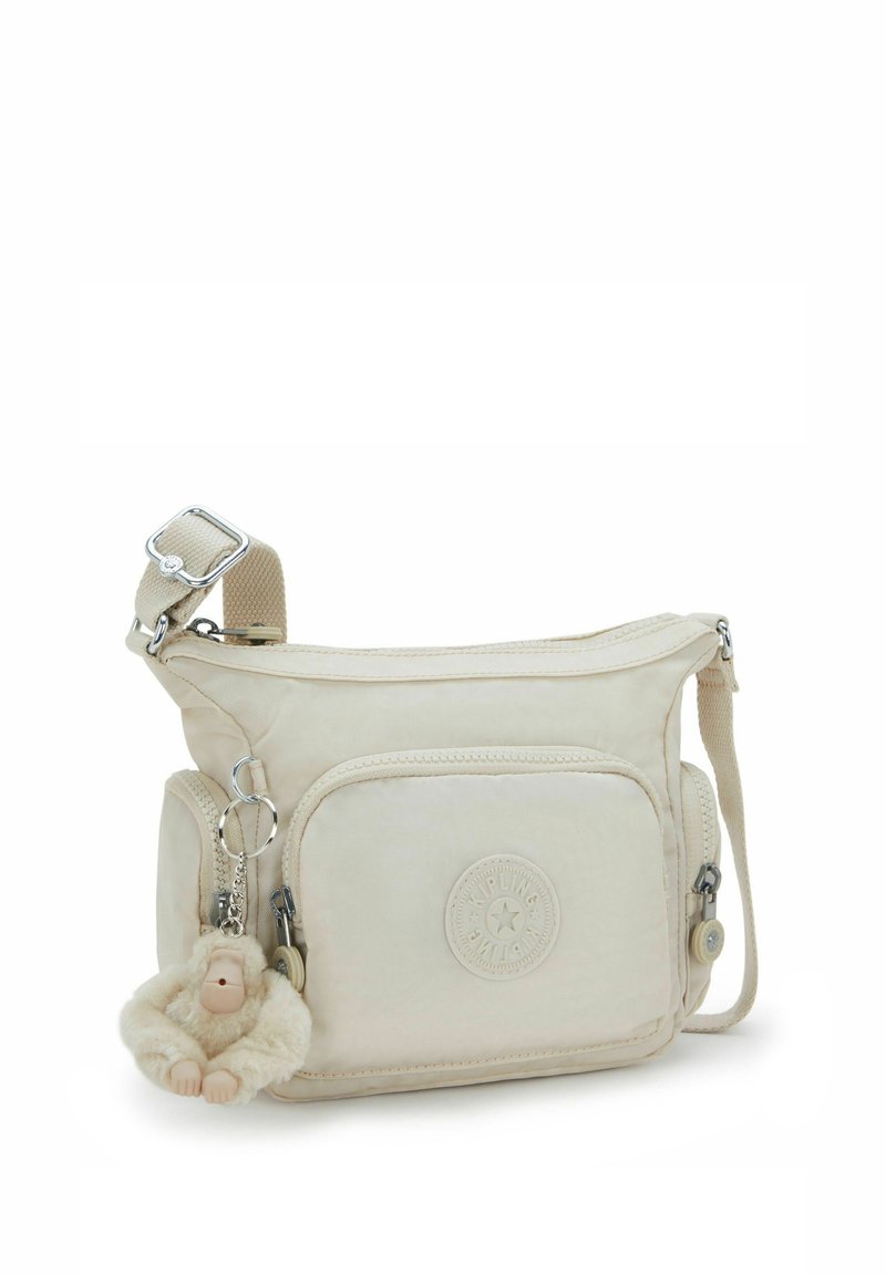 Beige Stoff Umhängetasche mit einer Frontreißverschluss-Tasche, einem Logo-Patch und einem Affenschlüsselanhänger. Mit verstellbarem Schulterriemen und glatter Textur.
