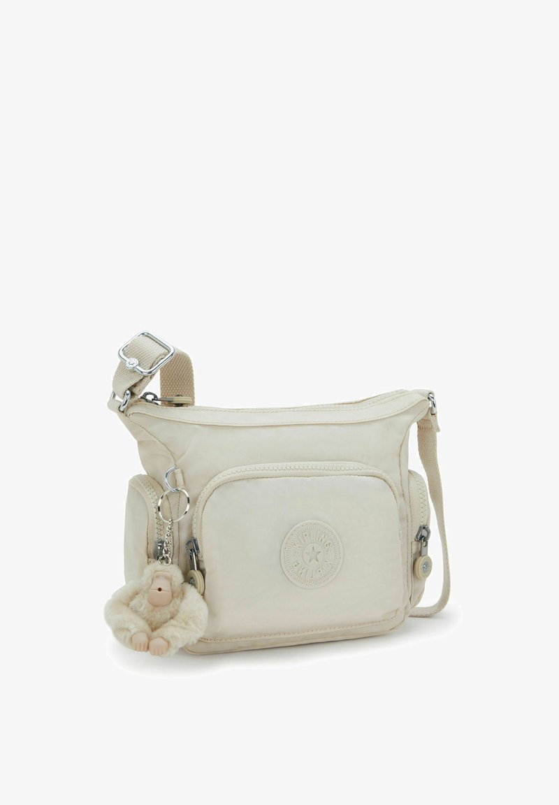 Beige fabric crossbody bag with a front zipper pocket, logo patch, and a monkey keychain. Features an adjustable strap and smooth texture.