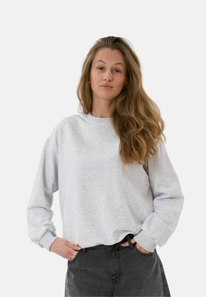 Vrouw met lang golvend haar die een lichtgrijze trui en donkere spijkerbroek draagt, staat met handen in de zakken tegen een effen achtergrond.