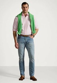 Man in light pink button-up shirt with green sweater draped over shoulders, blue jeans, brown belt, watch, and brown loafers standing on plain background.
