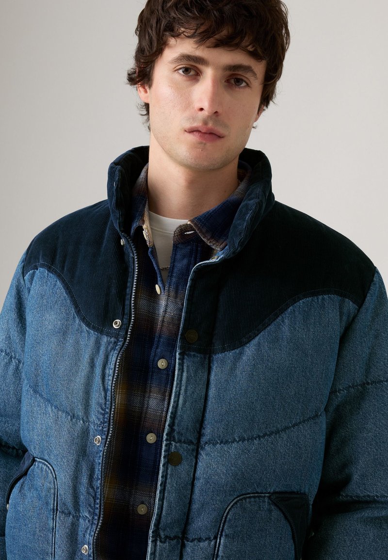 Denim pufferjakke med et gradient af blå nuancer, kordfløjlsdetaljer og knaplukning, lagt over en rutet flannel skjorte.