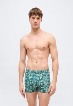 Fiatal férfi rövid barna hajjal, felsőtest nélkül, türkiz mintás úszónadrágban áll egy egyszerű, világos háttér előtt.