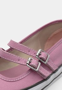 Sapatilha de lona rosa com duas alças ajustáveis e fivelas prateadas. Apresenta uma ponta arredondada e sola de borracha branca com contornos pretos.