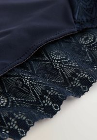 Sötétkék fürdőruha, sima, matt anyagból készült, amely a tetején található, és bonyolult csipke díszítéssel rendelkezik az alján. Geometrikus minták és fodros szélek jellemzik.