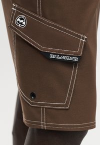 Καφέ cargo σορτς με λευκά ράμματα, τσέπη με καπάκι, μαύρο κουμπί και ετικέτα με το λογότυπο Billabong στο πλάι κοντά στο τελείωμα.