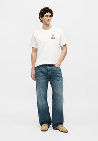 Ein Mann steht und trägt ein weißes PS Paul Smith T-Shirt, blaue weite Jeans und beige Loafers, mit einer Hand in der Tasche.