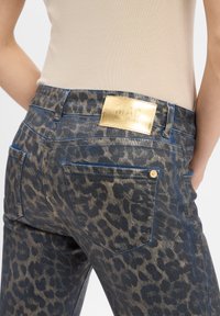 Jean en imprimé léopard avec denim bleu foncé, quincaillerie en or, et une étiquette dorée bien visible au dos de la ceinture. Comprend deux poches arrière.