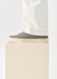 Γκρι σουέτ αθλητικά παπούτσια με επίπεδες μπεζ καουτσούκ σόλες και στρογγυλές κορδόνες, τοποθετημένα σε μια ανοιχτόχρωμη ξύλινη βάση. Λευκά βαμβακερά παντελόνια μερικώς ορατά.