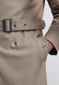 Trench-coat beige avec ceinture, grands boutons, poche latérale avec une main partiellement insérée et une petite étiquette en cuir.