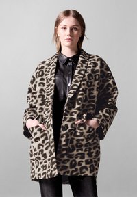 Leopardikuosis takki, joka on valmistettu pehmeästä kankaasta, avonaisella etuosalla, leveillä hihoilla ja kahdella etutaskulla, mustan paidan päällä.