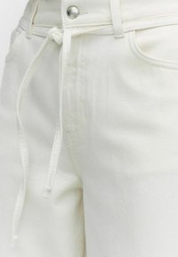 Vita denimbyxor har en dragsko i midjan, framfickor och en slät textur. Tyget är något strukturerat med en ren finish.