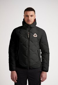 Svart puffjacka med hög krage, kviltad textur och sidofickor. Har en logotyp i en orange och vit triangel på bröstet.