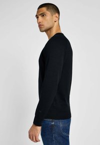 Zwarte trui met een ronde hals, lange mouwen en ribgebreide onderrand. Het model draagt deze met een blauwe spijkerbroek, waardoor een aansluitende silhoutte zichtbaar is.