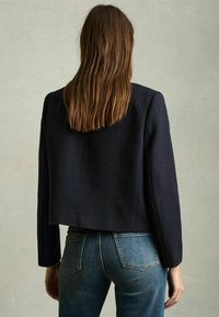 Tumes teal cropped bleiser tekstureeritud kangast, pikkade varrukatega ja struktureeritud õlgadega, kombineeritud kitsaste siniste teksadega.