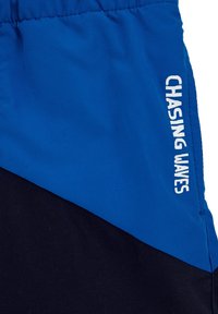 Mėlynos ir juodos sportinės šortai su minkšta tekstūra, turi baltą užrašą "CHASING WAVES" mėlynoje dalyje ir elastingą juosmenį.
