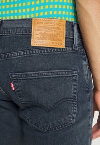 Vaqueros Levi's 502 de denim oscuro con un parche de cuero marrón que presenta detalles de la marca en relieve. Incluye una etiqueta roja en el bolsillo trasero.