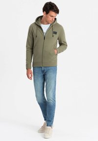 Canguro verde oliva con cremallera, capucha, bolsillos frontales y etiqueta de logo. Combinado con jeans azul claro y zapatillas beige.