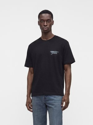 Uomo che indossa una t-shirt nera con una piccola scritta "Community Garden" sul petto e jeans blu, in piedi contro uno sfondo bianco.