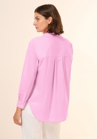 Camicia rosa chiaro a maniche lunghe con colletto, caratterizzata da una piega sul retro e polsini con bottoni, abbinata a pantaloni bianchi. Texture liscia e morbida.