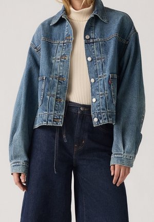 Femme portant une veste en jean bleue oversize par-dessus un pull à col roulé côtelé crème et un jean large foncé, debout devant un fond uni.