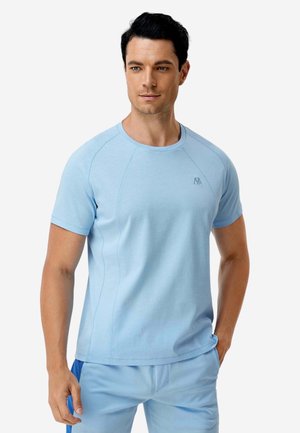 Uomo che indossa una maglietta azzurra a maniche corte con collo rotondo e pantaloni abbinati, in piedi con una mano in tasca davanti a uno sfondo neutro.