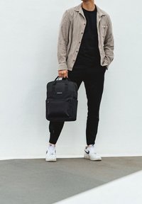 Mężczyzna ubrany w czarne spodnie, białe trampki i beżową kurtkę, trzymający czarny plecak za uchwyty na tle białej ściany.