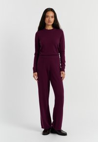 Koordiniertes Set aus lila Strickpullover und weit geschnittenen Hosen, das über einen gerippten Kragen und Nahtdetails verfügt und mit dunklen Schuhen kombiniert ist.