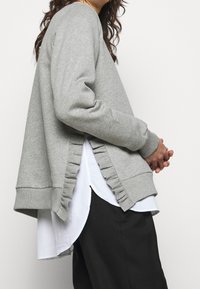 Grauer Sweatshirt mit strukturiertem Rüschendetail an den Seiten, über einem weißen Hemd getragen. Hat lange Ärmel und einen lässigen Schnitt.
