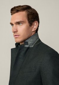 Mörk texturerad kappa med hög krage som har en grå tygaccent. Designen inkluderar en enkel framknäppning och sidofickor.