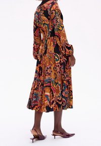 Robe florale multicolore avec de longues manches bouffantes, taille cintrée et jupe fluide. Fond sombre avec des motifs vibrants. Associée à des chaussures violettes.