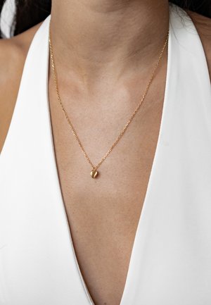 Goldfarbene Halskette mit einer zarten Kette und einem kleinen herzförmigen Anhänger, getragen über einem weißen Kleidungsstück. Glatte Textur und glänzende Oberfläche.