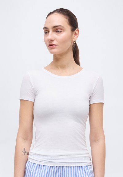 Jeune femme aux cheveux bruns attachés en queue de cheval, portant une chemise blanche cintrée à manches courtes et un pantalon rayé bleu et blanc, regardant vers la gauche.