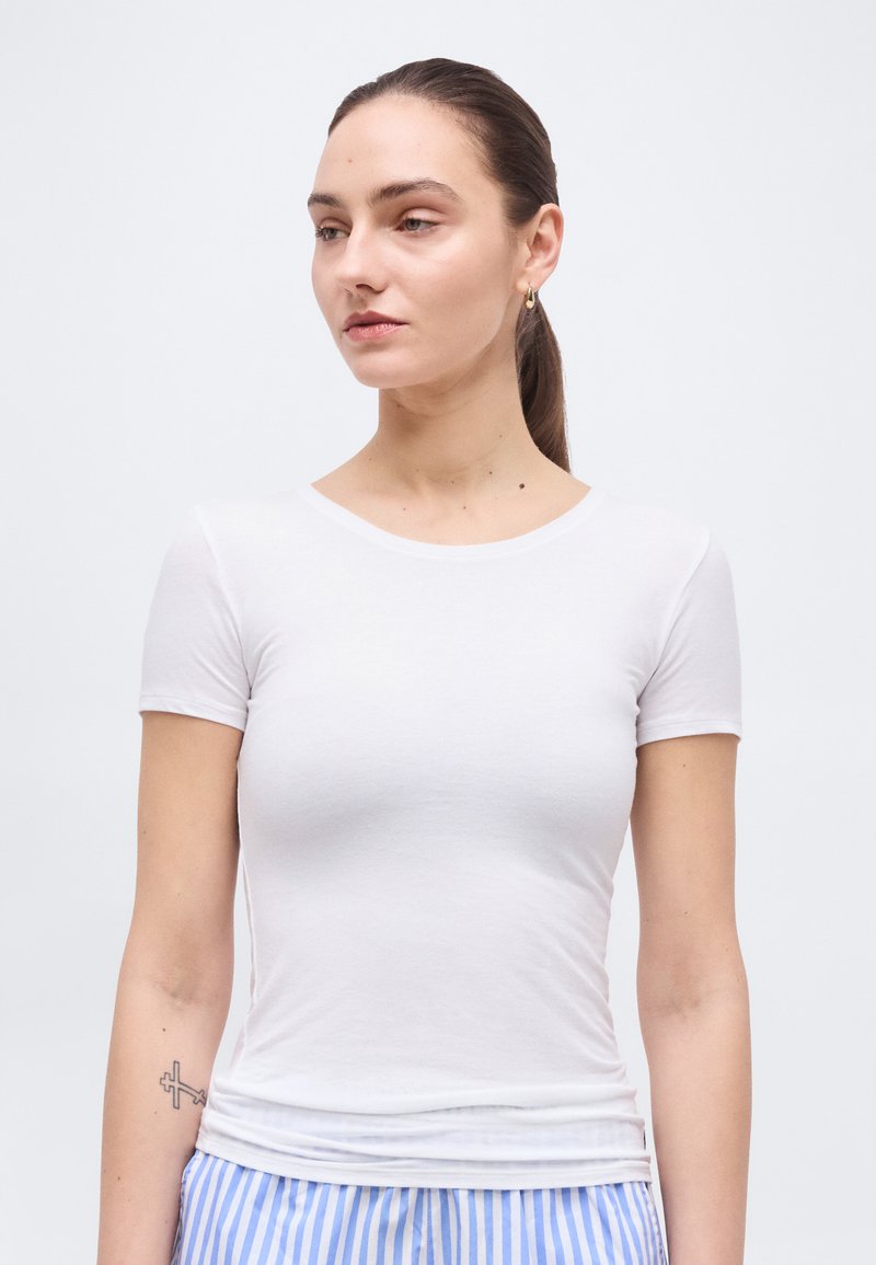 Νεαρή γυναίκα με καστανά μαλλιά σε αλογοουρά, φορόντας ένα λευκό εφαρμοστό μπλουζάκι με κοντά μανίκια και μπλε και λευκή ριγέ παντελόνα, κοιτάζει προς τα αριστερά.