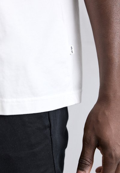 T-shirt de algodón blanco con una etiqueta lateral, con dobladillo recto y mangas cortas. Se muestra junto a un par de pantalones negros.