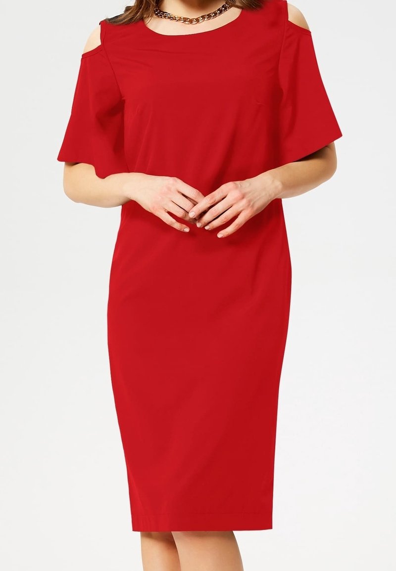 Femme portant une robe rouge longueur genou avec des découpes aux épaules et un collier chaîne doré, mains jointes à la taille.