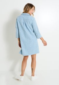 Robe en denim bleu clair avec broderies florales, manches trois-quarts, un col et une coupe décontractée. Portée avec des baskets blanches.