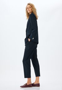 Marineblauwe op maat gemaakte blazer met een gestructureerde pasvorm, twee voorzakken en zilveren knopen. Gecombineerd met bijpassende broek en bordeauxrode instappers.