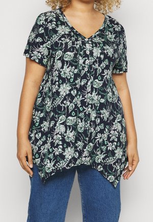 Personne aux cheveux bouclés portant un haut à manches courtes bleu marine à motifs floraux avec des boutons et un ourlet asymétrique, associé à un jean bleu.