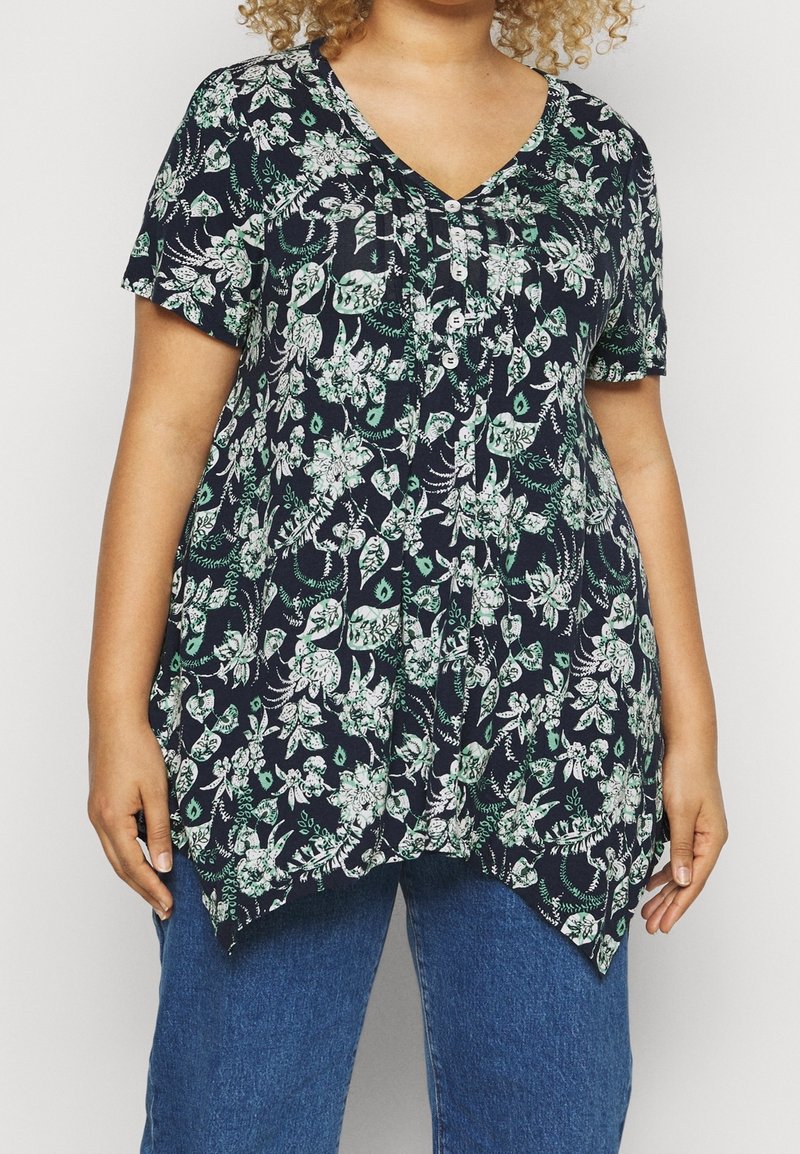 Persona con cabello rizado, vistiendo una blusa floral de manga corta en azul marino con botones y un dobladillo asimétrico, combinada con jeans azules.