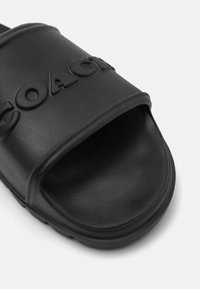 Black slide sandal made from textured synthetic material with a wide strap featuring embossed "COACH" branding. Open toe design.