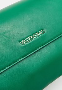 Zelená kožená kabelka Valentino s viditelným texturovaným povrchem a výrazným stříbrným logem značky na předním klopě.