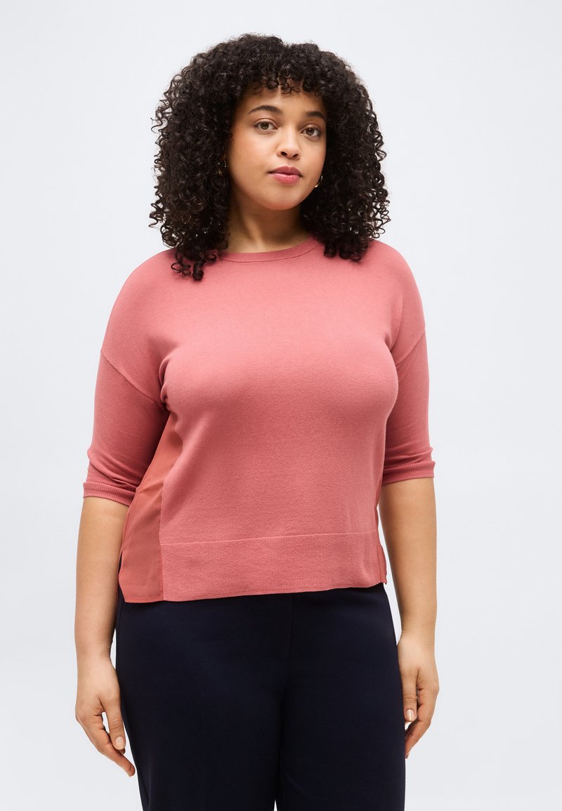 Γυναίκα με σγουρά μαλλιά που φοράει ροζ μπλουζάκι με κοντά μανίκια και σκούρο παντελόνι, στέκεται μπροστά σε απλό φωτεινό γκρι φόντο.