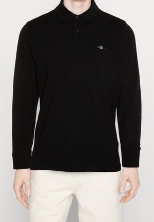 Mann trägt ein schwarzes Langarm-Poloshirt mit kleinem gesticktem Logo auf der linken Brustseite und hellbeige Hose.