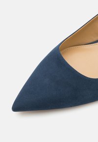 Chaussure plate en daim bleu marine à bout pointu avec doublure intérieure beige sur un fond clair uni.