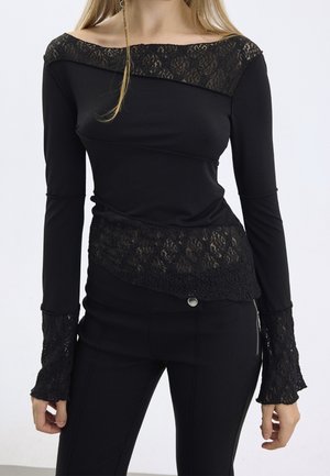 Vrouw draagt een zwarte top met lange mouwen en kantdetails bij de halslijn, taille en manchetten, gecombineerd met een zwarte broek, staand tegen een witte achtergrond.