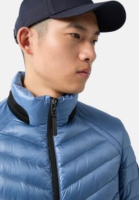 Blue insulated jacket with a high collar, diagonal quilted pattern, zip closure, and black mesh accents. Complemented by a dark cap.