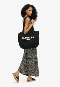 Schwarze gewebte Tragetasche mit dem Text "SUPERDRY IBIZA" in Weiß. Das Modell trägt ein gemustertes Maxi-Kleid in Schwarz und Weiß sowie Sandalen.
