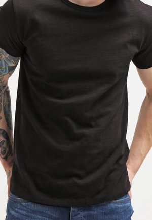 Mężczyzna w prostym czarnym t-shircie i niebieskich jeansach, z rękawem tatuażu na lewej ręce, stojący z rękami w kieszeniach.